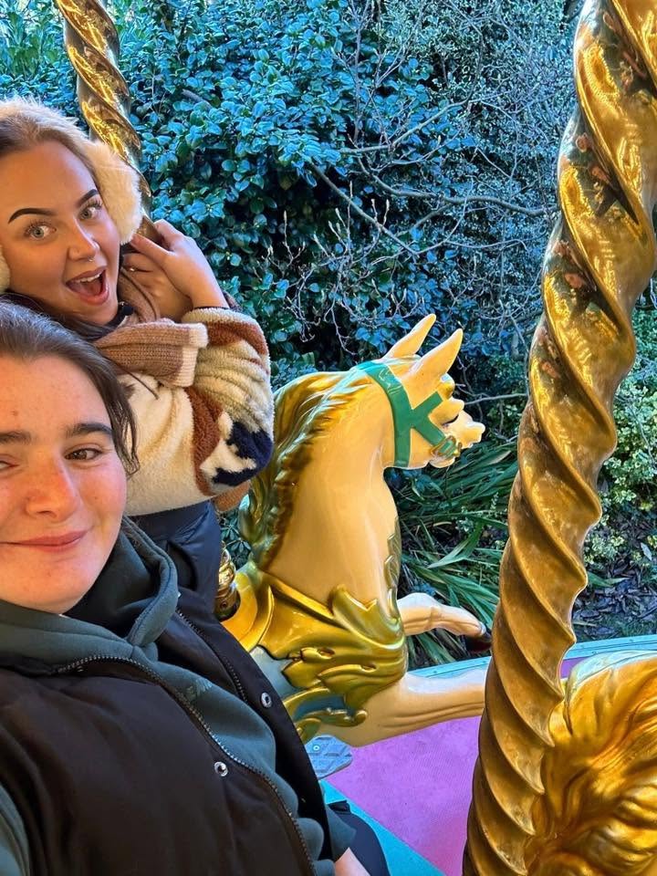 Two young women smiling at the camera beside a cream and gold carousel horse, with a twisted gold pole and a green hedge behind them