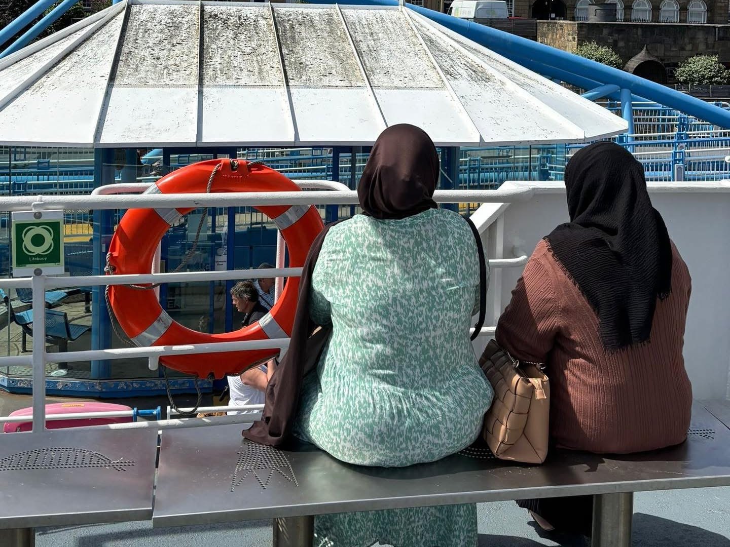 Two women sitting on the deck of a ferry, looking out towards buildings on the shore