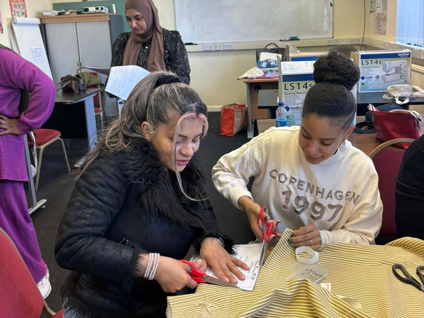 Two women cutting a length of striped fabric with scissors at a classroom table