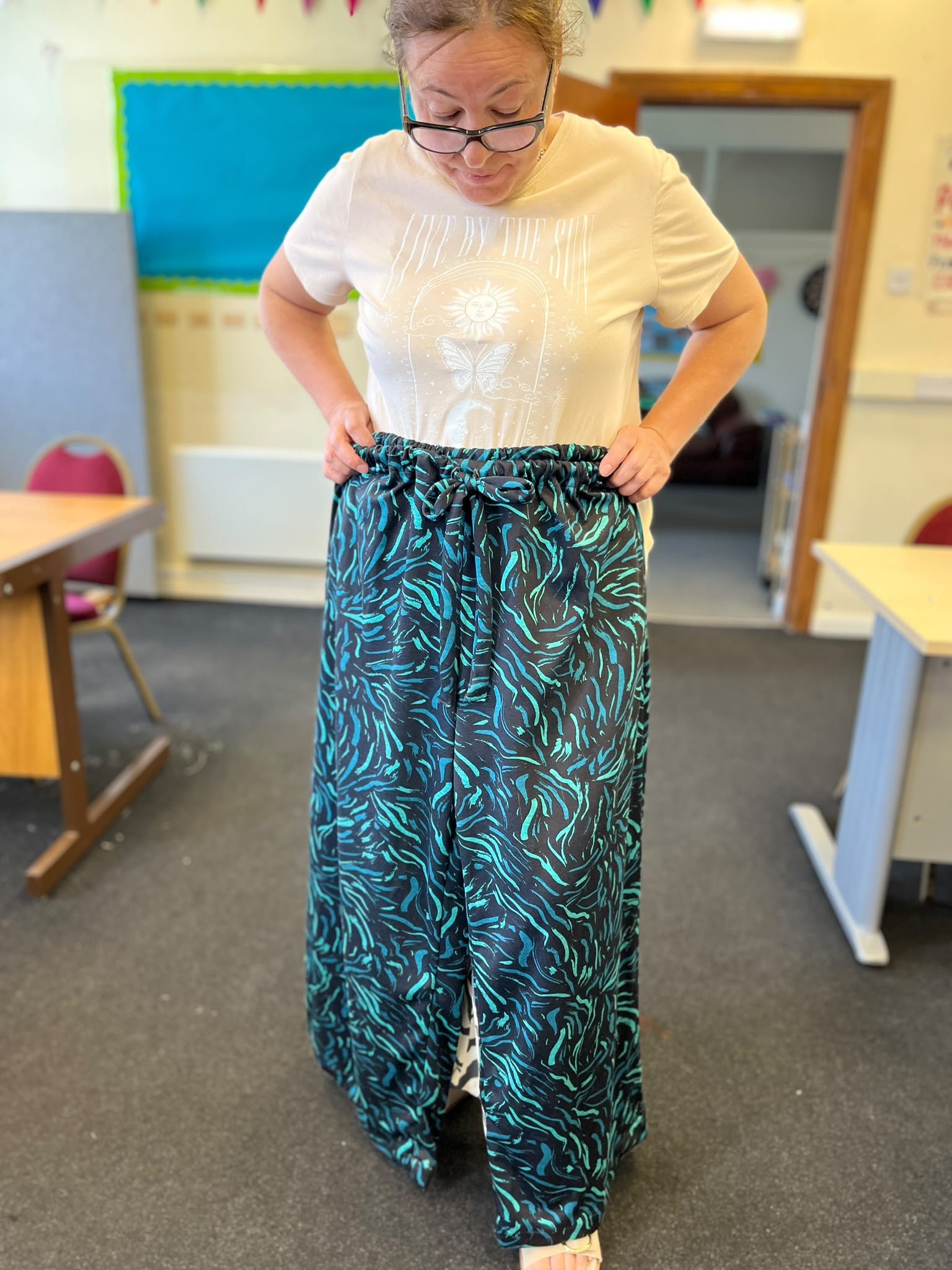 A woman standing in a classroom wearing wide green and black patterned trousers, holding the waistband