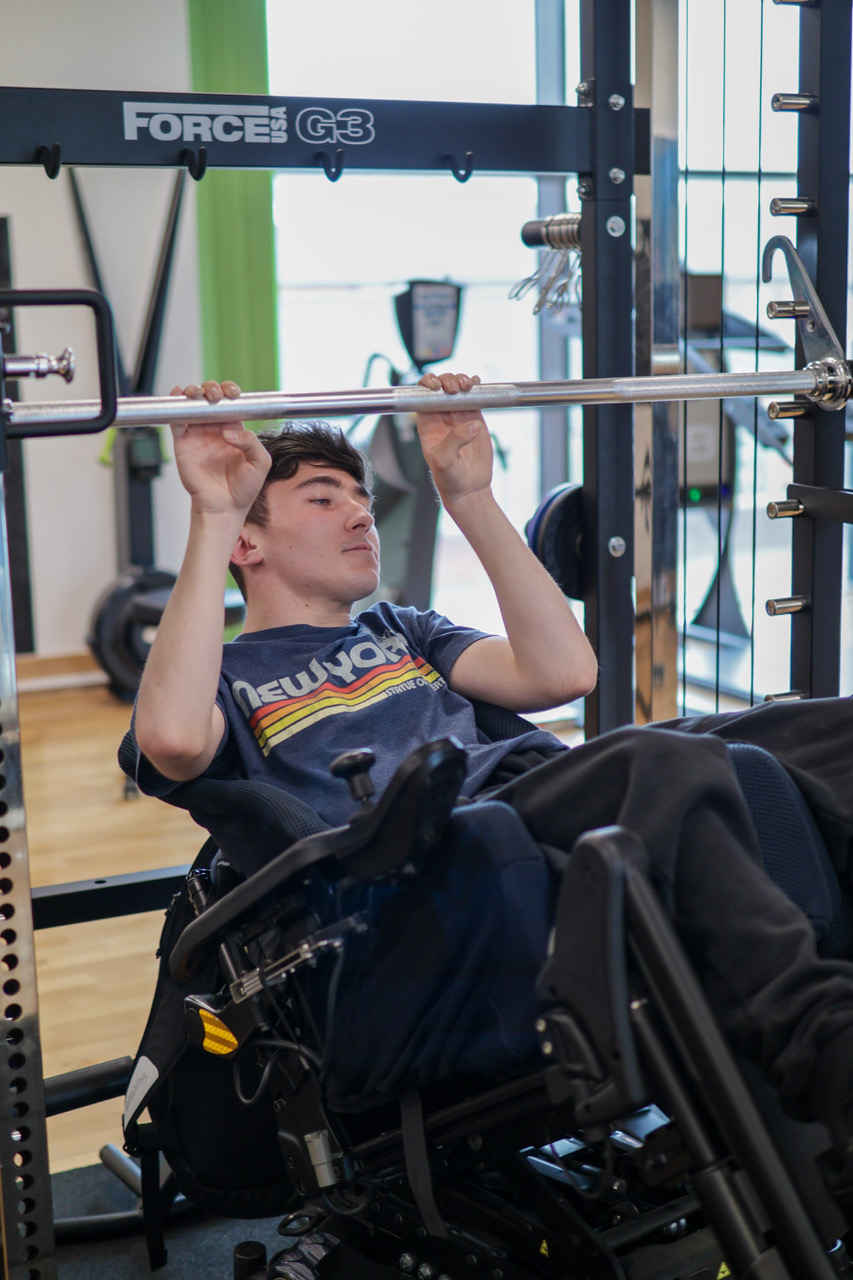 A young man in a powered wheelchair gripping a barbell with both hands on a gym rack, with gym equipment behind him