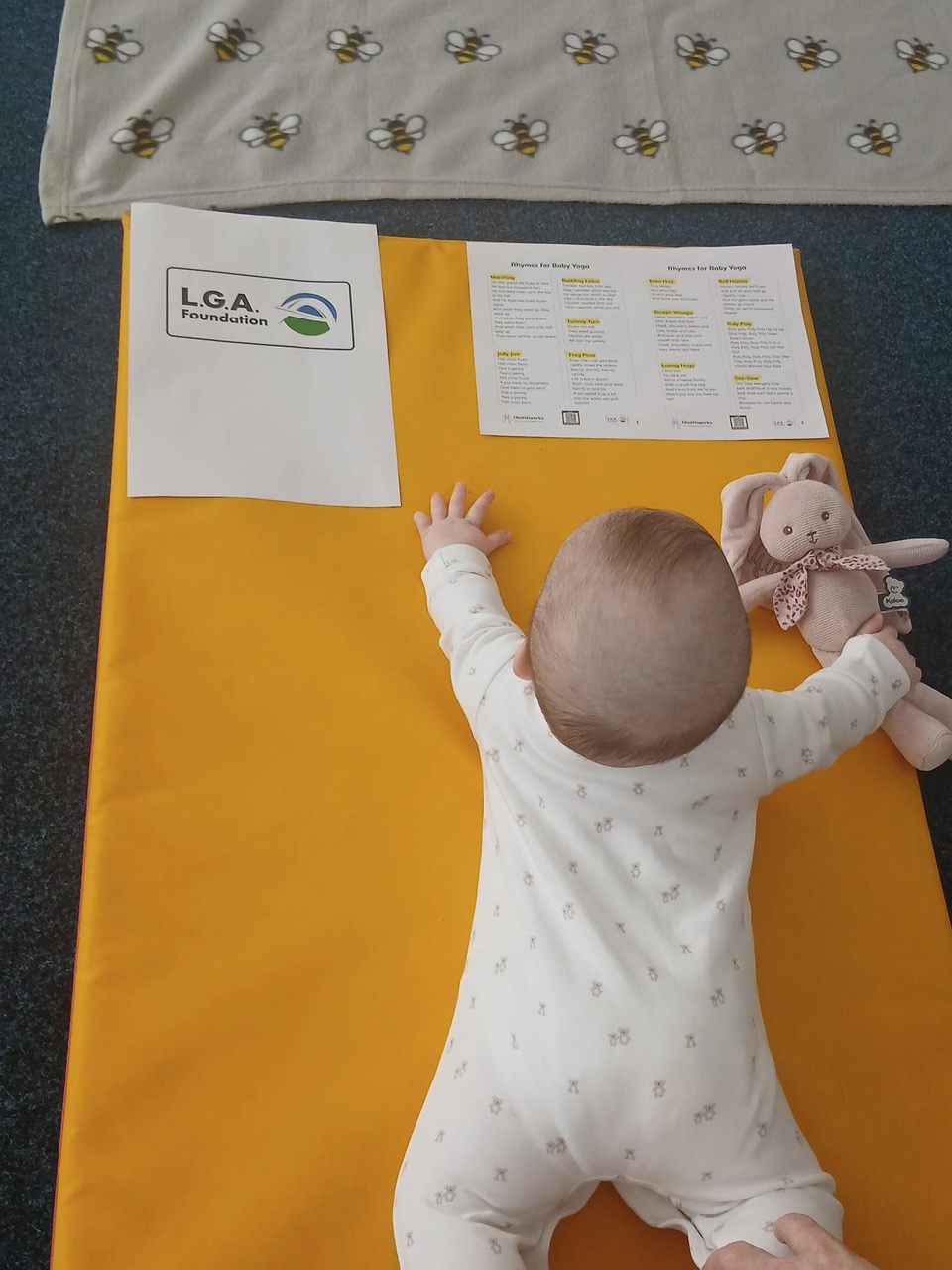 A baby in a white sleepsuit lying on a yellow mat, reaching towards a printed sheet of baby yoga rhymes, with a soft toy rabbit alongside