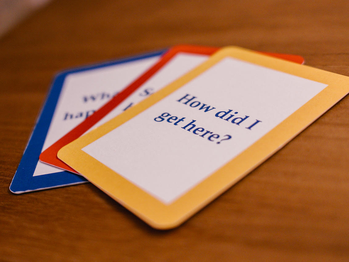 Three coloured question cards fanned out on a wooden table, the top one reading How did I get here?