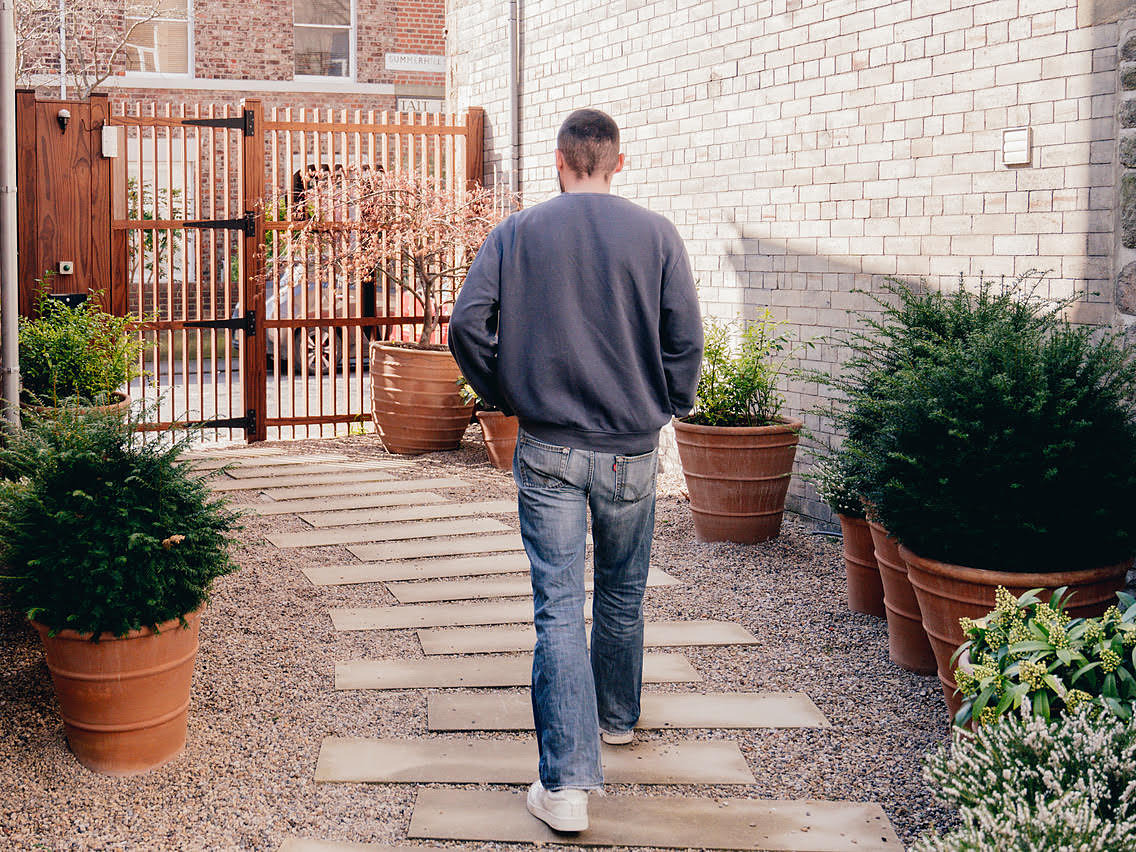 A man walking along a stone path through a planted courtyard towards a wooden gate