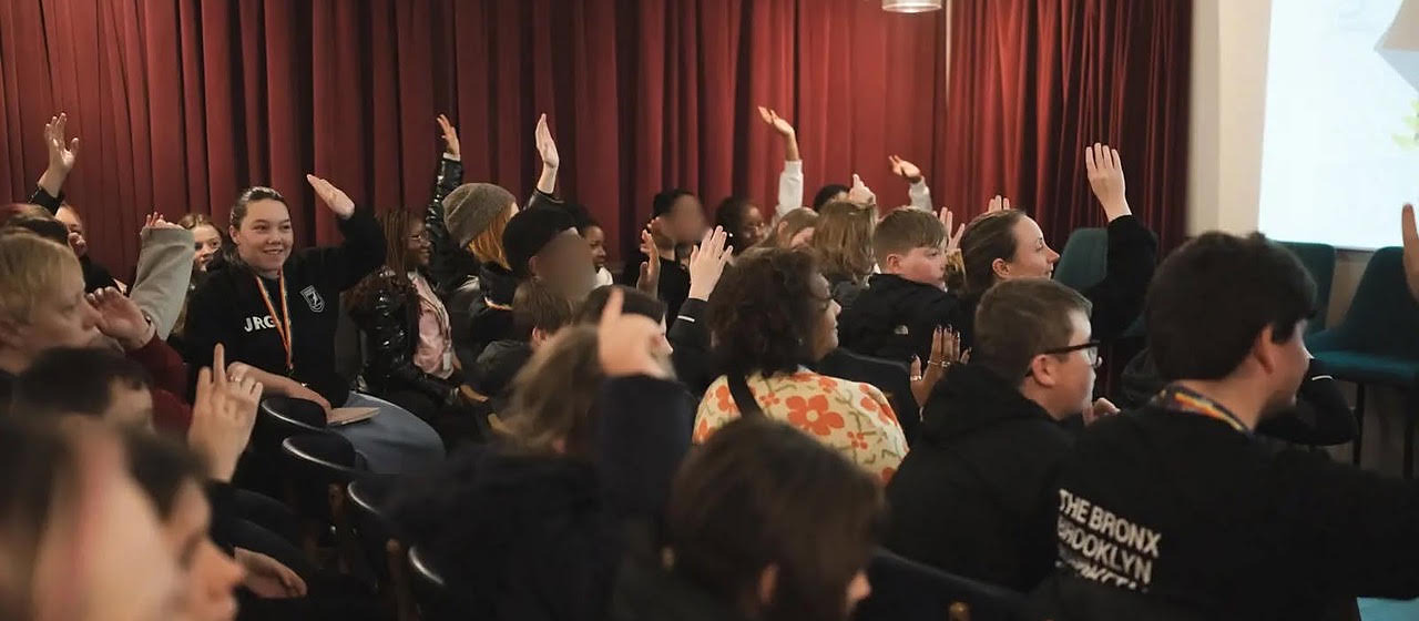 A hall full of young people seated with their hands raised