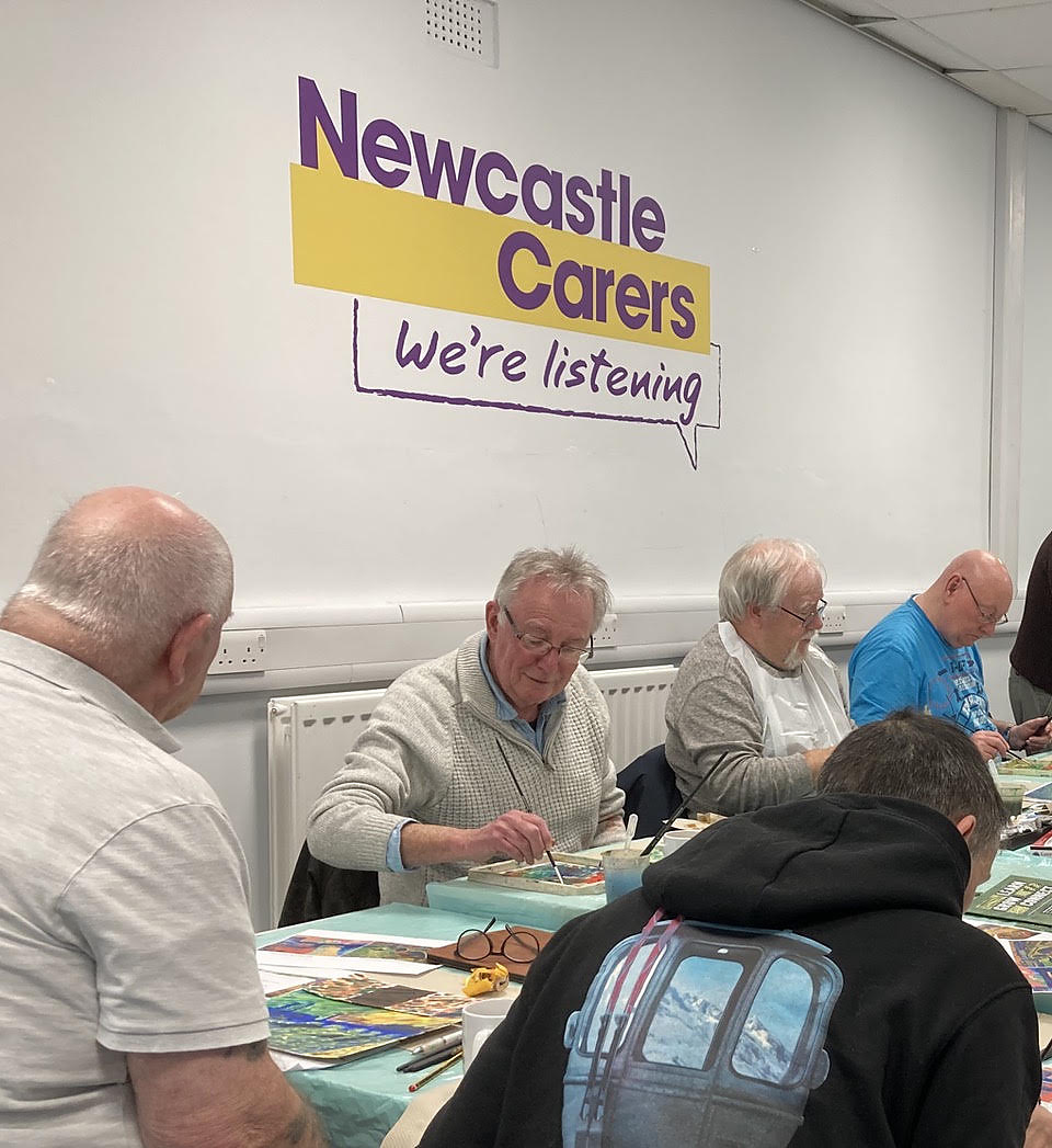 People sitting around tables at a craft session beneath a Newcastle Carers banner reading We are listening
