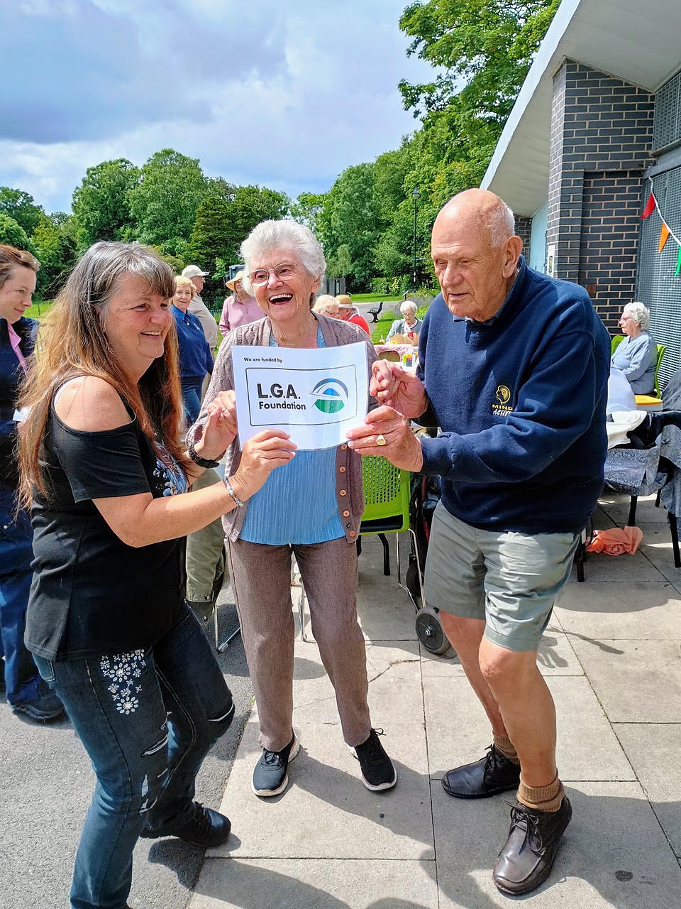 Two people presenting a printed LGA Foundation certificate to a woman at an outdoor event