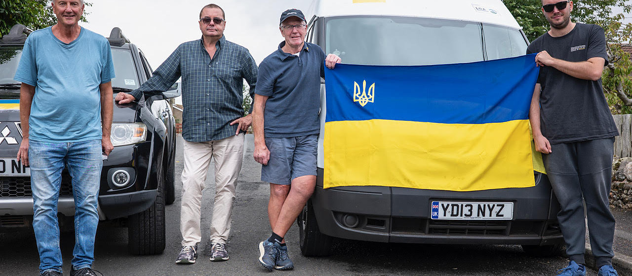 Four men standing beside a van with a Ukrainian flag draped across its back doors
