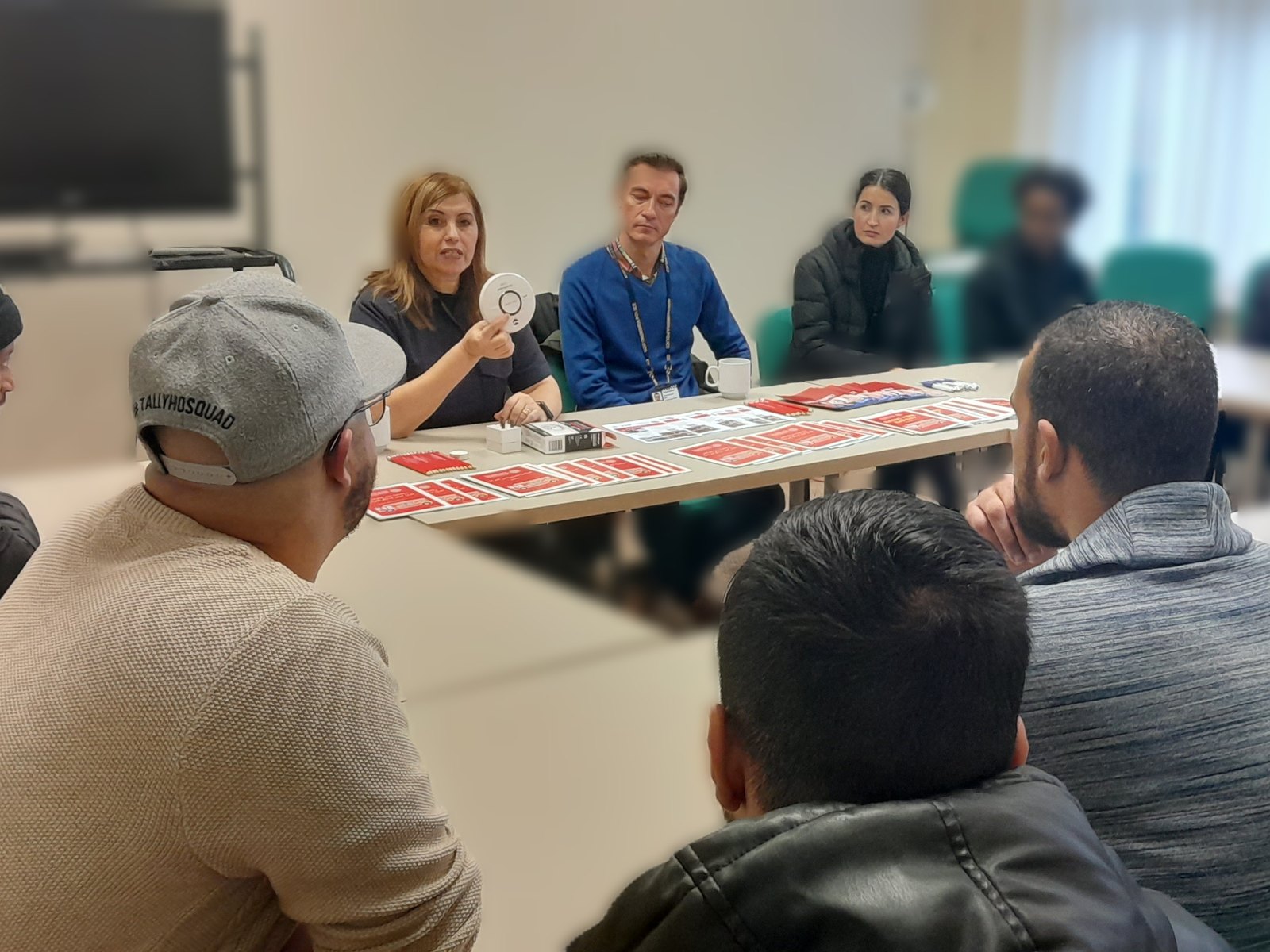 A group of people seated around a long table in a meeting room, listening to a woman holding up a card