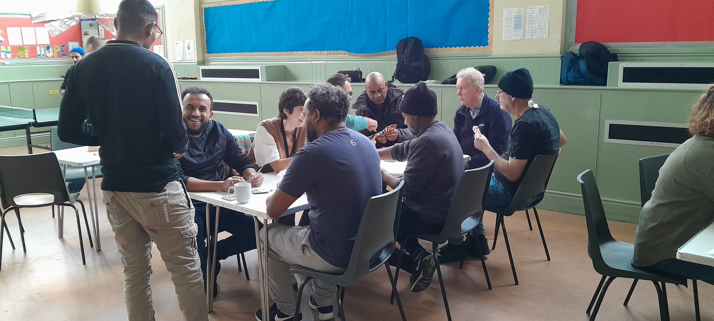 A group of men sitting around tables in a community hall, talking and playing cards, with one man standing beside them