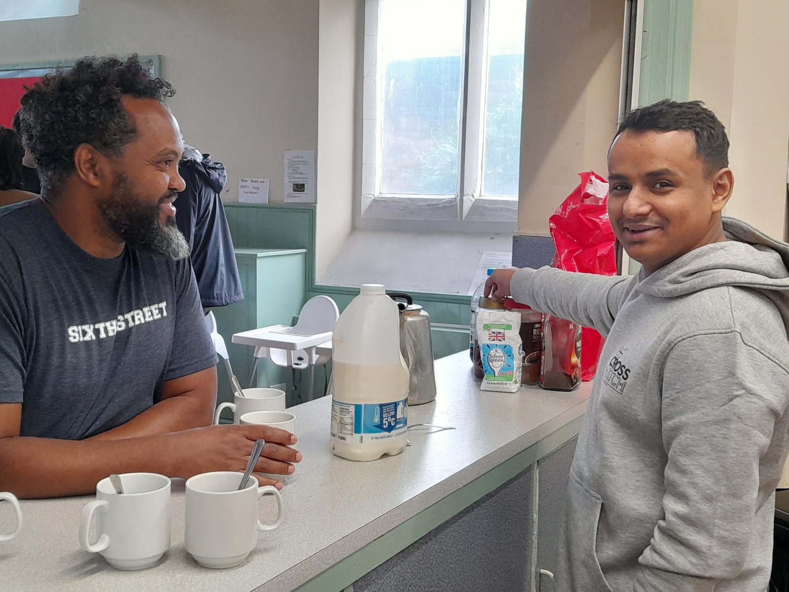 Two men smiling at a kitchen counter with mugs and a carton of milk